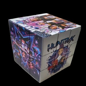 May include: A 3x3 puzzle cube featuring a custom graphic design with the text HUNTRX. The cube displays colourful illustrations of characters, blue neon-style stars, and abstract shapes across its rotating segments on a white background.