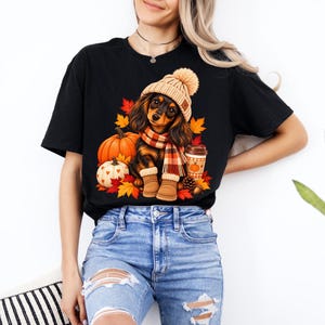 Può includere: Una t-shirt nera con la grafica di un bassotto a pelo lungo che indossa un berretto di lana beige, una sciarpa a quadri e stivali color cuoio. Il cane è circondato da zucche arancioni e bianche, foglie autunnali e un bicchiere di caffè.