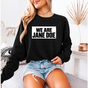 May include: A black long-sleeve sweatshirt featuring a white rectangular graphic on the chest with the bold black text WE ARE JANE DOE printed in a sans-serif font. The garment has a crew neckline and a standard fit with long sleeves.