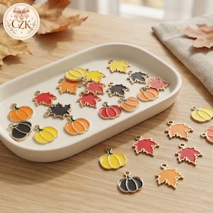 May include: A collection of small metal charms shaped like autumn leaves and pumpkins, featuring colourful enamel fill in shades of red, orange, yellow, and black. Each charm includes a small loop at the top for attachment to jewellery or crafts.