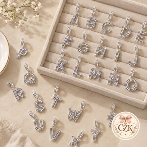 May include: A collection of silver-toned alphabet letter charms, each featuring a pavé-style surface with small white stones. The charms are arranged in a light-colored jewelry tray and scattered on a surface, showcasing letters A through Z.