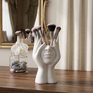Peut inclure: Un porte-pinceaux en céramique blanc mat représentant deux mains entourant un visage, rempli de divers pinceaux de maquillage. La sculpture présente des traits faciaux détaillés, incluant un nez et des lèvres, servant d'organisateur fonctionnel.