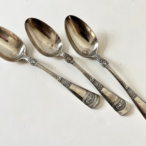 May include: Three silver spoons with ornate handles and engraved initials. The spoons are arranged in a row on a white surface.