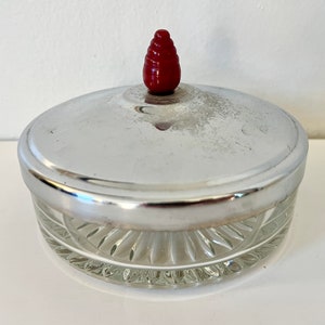 Könnte beinhalten: Ein klares Glasgefäß mit einem silbernen Deckel und einem roten Plastikknopf oben. Das Gefäß hat ein dekoratives, geriffeltes Design.