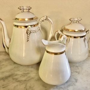 May include: A white porcelain tea set with gold trim. The set includes a teapot, creamer, and sugar bowl. Each piece features a rope design and a gold band around the top.