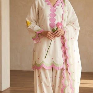 May include: An off-white salwar kameez set featuring a V-neckline with pink heart-shaped embroidery, scalloped edges in pink and light green, small pink heart patterns throughout, a yellow sun motif on the sleeve, and a matching scalloped dupatta.