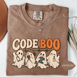 May include: A brown short-sleeve t-shirt with a graphic print featuring four ghosts dressed as medical professionals. The text reads CODE BOO. The ghosts are depicted with a stethoscope, a coffee cup, a clipboard, and a syringe. The shirt has a Comfort Colors label.