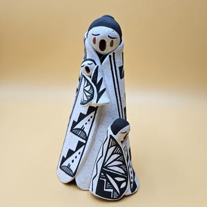 May include: A ceramic figurine featuring a large figure wrapped in a white blanket with black geometric patterns, holding a smaller figure in a matching blanket, with a third small figure standing at the base, all with black hair and painted faces.