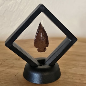 Può includere: Una punta di freccia marrone esposta in una teca nera a forma di diamante con una base nera.