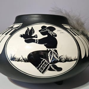 Op de afbeelding: Zwart-witte keramische pot met een Indiaans ontwerp. De pot heeft een centraal ovaal ontwerp met een figuur die een vogel vasthoudt. Er zijn veren aan de bovenkant van de pot bevestigd.