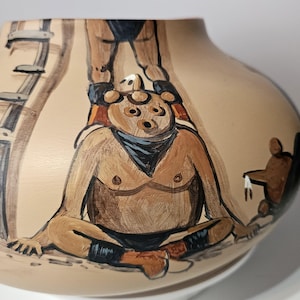 Puede incluir: Un jarrón de cerámica beige con un diseño pintado a mano que presenta figuras de estilo suroeste. Las figuras están representadas en tonos de marrón, negro y azul. Una figura está sentada y otra está de pie encima. También hay una escalera pintada en el jarrón.