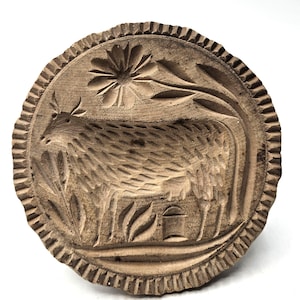 Pode incluir: Um molde de manteiga redondo de madeira com um design esculpido de uma vaca em um campo, com uma flor acima e um pequeno balde perto dos pés. A peça circular tem uma borda texturizada e festonada com um acabamento de madeira clara natural.