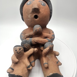 May include: A brown ceramic figurine of a person wearing a hat, holding three smaller figures. The figurine has black detailing on the hat, arms, and legs. The figures have simple faces and are arranged around the central figure.