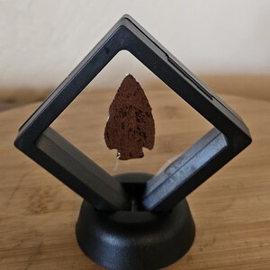 May include: A brown arrowhead displayed in a black diamond-shaped display case with a black base.