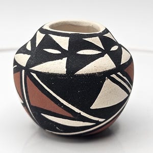 Puede incluir: Una pequeña olla redonda de cerámica con un diseño geométrico. La olla presenta un patrón negro, blanco y marrón rojizo. El borde es de color beige claro. El diseño es simétrico y cubre toda la superficie de la olla.