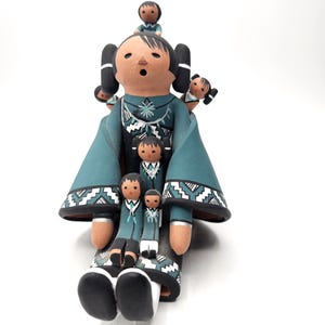 May include: A clay storyteller figurine featuring a large central figure with black hair and a teal robe, surrounded by five smaller figures perched on the head, shoulders, and lap. The clothing is decorated with white and black geometric patterns.