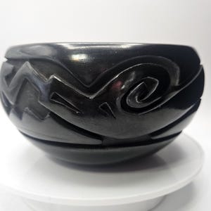 Może przedstawiać: Czarna, okrągła ceramiczna miska z rzeźbionymi geometrycznymi wzorami. Miska ma zygzakowate i wirujące wzory na zewnątrz. Miska jest ustawiona na białym tle, podkreślając jej ciemny kolor i skomplikowane detale.