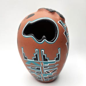 Puede incluir: Un pequeño jarrón de cerámica de terracota pintado a mano con un diseño de oso. El jarrón está decorado con patrones geométricos en negro, turquesa y blanco.