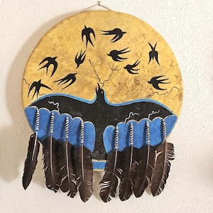 May include: A circular drum with a tan, textured surface featuring a black painted bird with wings spread over a blue wave-like pattern. Above the bird, several black swallow silhouettes are painted. Eight dark brown feathers hang from the bottom.