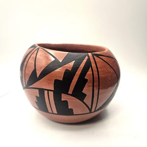 Puede incluir: Un cuenco de cerámica redondo, color terracota, con un diseño geométrico negro. El cuenco tiene una abertura ancha y un acabado liso y brillante. El diseño presenta formas y líneas angulares, creando un contraste llamativo.