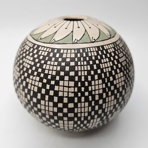 Może przedstawiać: Sferyczny ceramiczny wazon z czarno-białym geometrycznym wzorem kratki pokrywającym korpus. Górna część przedstawia okrągły pas z jasnozielonym tłem i serią białych, przypominających płatki kształtów z czarnym obrysem.