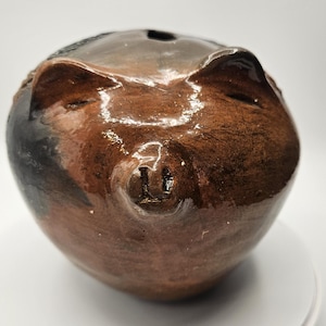 May include: A brown ceramic piggy bank with a glossy finish. The bank has a slot on top and features a pig's face with ears and a snout. The colors range from dark brown to black.