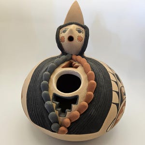 May include: A hand-painted ceramic pot with a stylized human figure on top. The pot is decorated with black, brown, and white geometric patterns. The figure has a pointed hat and a round face with black eyes and a small mouth.