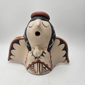 May include: A ceramic angel figurine with a cream-colored body, black hair, and a brown halo. The angel has closed eyes and folded hands, with black and brown geometric patterns on the wings and clothing. The figurine has a hole in the mouth.