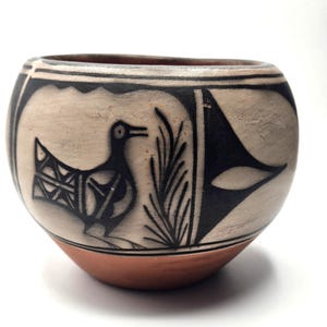 May include: A small, round, white ceramic pot with black painted designs. The pot features a bird, a plant, and geometric shapes.