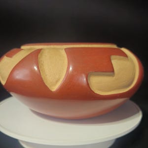 Può includere: Una ciotola artigianale in ceramica, marrone rossiccio, con un design unico. La ciotola presenta una serie di forme curve color beige chiaro scolpite all'esterno. La ciotola è rotonda con un'ampia apertura e poggia su una superficie bianca.