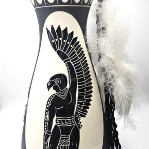 Op de afbeelding: Een zwart-crème keramische vaas met een decoratief ontwerp met een figuur met vogelachtige kenmerken en uitgespreide vleugels. De vaas heeft een uitlopende bovenkant en is versierd met witte veren.