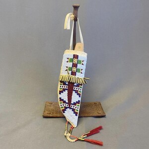 May include: A beaded knife sheath with a white background and geometric patterns in red, green, blue, and yellow. The sheath has a fringe of gold-colored beads and red tassels. It is suspended from a wooden stand.