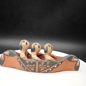 May include: A handcrafted ceramic canoe with three stylized figures. The canoe is a warm terracotta color, accented with black and white geometric patterns. The figures have tan bodies and black hair.