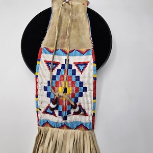 May include: A tan leather pouch featuring a geometric beaded pattern in white, blue, red, and yellow. The pouch includes a long leather drawstring with two brown and white beads, and a long fringe at the bottom made of thin tan leather strips.