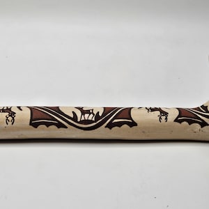 May include: A long, white ceramic pipe with a brown design of stylized deer and geometric patterns. The pipe has a wide, flat base and a round, open end.