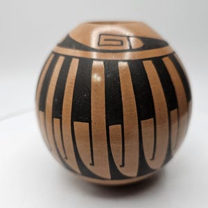 May include: A round, tan-colored ceramic vase with a black geometric design. The vase features vertical black stripes and a spiral design near the opening. The vase has a smooth, glossy finish.