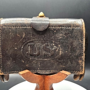 Op de afbeelding: Een vintage, donkerbruine leren portemonnee met de letters "US" in reliëf in een ovaal. De portemonnee heeft een klepsluiting met een messingkleurige knop en wordt op een houten standaard getoond. Het leer vertoont tekenen van slijtage.