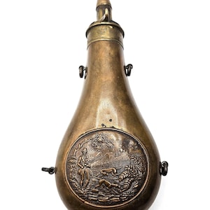 Peut inclure: Une poire à poudre en laiton vintage en forme de goutte avec un bec étroit au sommet. Le corps présente un relief circulaire embossé représentant un chasseur tenant un fusil accompagné de deux chiens de chasse dans un paysage rural.