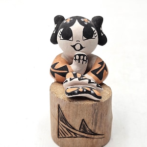 Może przedstawiać: Mała ceramiczna figurka stylizowanej postaci z czarnymi włosami, dużymi oczami i ubraniem w pomarańczowo-czarne wzory, spoczywająca na cylindrycznej drewnianej podstawie z wygrawerowanym geometrycznym wzorem trójkąta i linii.