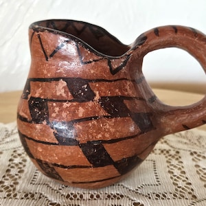 May include: A small, brown clay pitcher with a black geometric design. The pitcher has a rounded body and a handle.