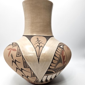 Puede incluir: Un jarrón de cerámica color canela con un cuerpo ancho y redondeado y un cuello estrecho. La superficie presenta patrones geométricos pintados a mano en negro, blanco y terracota, incluyendo motivos vegetales estilizados y formas triangulares.