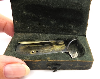 Puede incluir: Una navaja de bolsillo antigua de latón y acero en un pequeño estuche forrado de terciopelo verde oscuro. La navaja está abierta, revelando una hoja y otras herramientas. El estuche es rectangular con un cierre de broche.