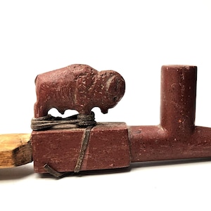 Puede incluir: Una pipa de piedra de color rojo parduzco tallada con una figura de bisonte esculpida y sujeta a la parte superior con un cordón de cuero fino. La pipa tiene un hornillo cilíndrico y una boquilla cónica, unida a un mango de madera clara.