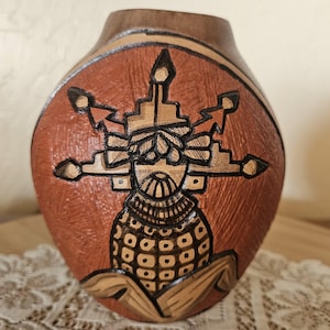 Puede incluir: Un jarrón tallado a mano, marrón y marrón rojizo, con un diseño intrincado. El motivo central es una figura estilizada con patrones geométricos. El jarrón tiene un cuerpo redondeado y un cuello estrecho, con una banda marrón claro en la parte superior.
