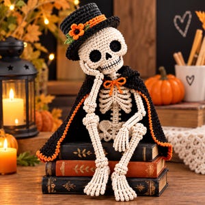 May include: A crocheted skeleton figurine wearing a black top hat with an orange flower, a black cape with an orange trim, and an orange bow tie. The skeleton is sitting on a stack of three books, resting its chin on one hand.