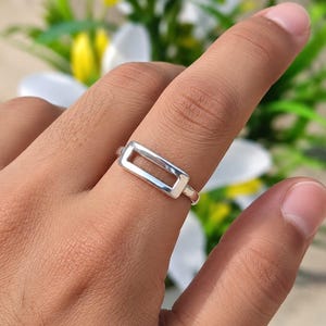 Könnte beinhalten: Ein polierter Silberring mit einem horizontalen, rechteckigen, offenen Rahmendesign. Das Band ist glatt und metallisch, umschließt den Finger mit einer klaren, geometrischen Form, die flach auf der Haut liegt und ein gleichmäßiges Silberfinish zeigt.