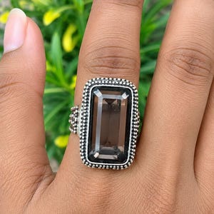 May include: A rectangular smoky quartz gemstone ring set in a silver band. The stone features a faceted emerald cut and is surrounded by a decorative beaded silver border. The metal band has a textured, dotted finish along the sides.