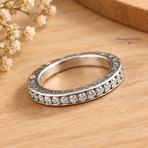 May include: A silver-toned eternity band featuring a continuous row of round, clear, faceted stones set along the front. The metal band displays an intricate, engraved floral or scrollwork pattern along the side, adding texture to the design.