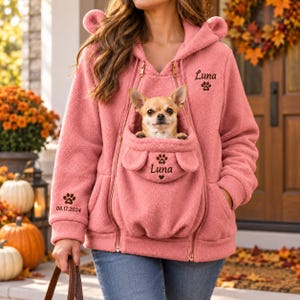 Könnte beinhalten: Ein altrosa Fleece-Kapuzenpullover mit einer Fronttasche mit Tierohren, entworfen für den Transport eines kleinen Hundes. Der Pullover zeigt den gestickten Text "Luna" mit Pfotenabdruck, ein zweites "Luna" auf der Tasche und "08.17.2024" am Ärmel.
