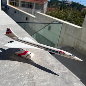 Könnte beinhalten: Ein weißes British Airways Concorde Modellflugzeug mit spitzer Nase, Deltaflügeln und einem Seitenleitwerk mit roten und blauen Akzenten. Der Rumpf trägt den Schriftzug British Airways und der Sockel ist mit dem Wort Concorde in Schwarz beschriftet.
